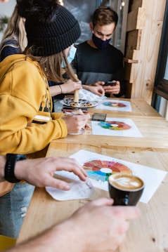 A group of people sitting at a wooden table engaged in an activity involving colorful charts. One person is holding a black coffee cup, while another person is drawing on a chart with a pencil. They are dressed casually, and some are wearing face masks, indicating it might be during the COVID-19 pandemic. The background includes some wood paneling and a window.