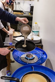 Two people in a kitchen are engaged in cooking. One person is pouring batter from a ladle into a frying pan over a blue gas stove. A bottle of sunflower seed oil, plates, and some cooking utensils are visible on the counter in the background. Both individuals are wearing aprons, and one of them is wearing a face mask.