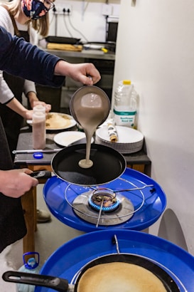 Two people in a kitchen are engaged in cooking. One person is pouring batter from a ladle into a frying pan over a blue gas stove. A bottle of sunflower seed oil, plates, and some cooking utensils are visible on the counter in the background. Both individuals are wearing aprons, and one of them is wearing a face mask.