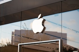 A large, white Apple logo is prominently displayed on the glass facade of a modern store. The building features reflective surfaces, capturing the sky and surrounding cityscape. The overall architecture is sleek and minimalistic.