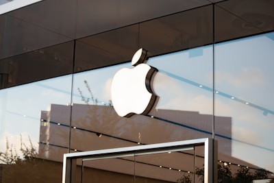 A large, white Apple logo is prominently displayed on the glass facade of a modern store. The building features reflective surfaces, capturing the sky and surrounding cityscape. The overall architecture is sleek and minimalistic.