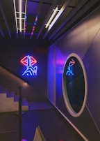 A modern interior space featuring a neon art installation on the wall, with a design of a hand and lips in vibrant blue and red colors. The area includes a circular mirror reflecting part of the neon light, and a set of stairs ascending to the left. Overhead lighting fixtures contribute to the ambiance, casting a soft glow.