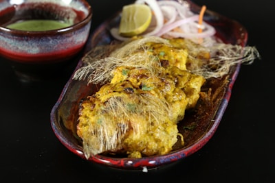 A dish featuring golden-brown grilled pieces of chicken, garnished with thin strands that resemble fried vermicelli, placed in a decorative oval plate. Accompanying the dish is a small bowl of green sauce, a wedge of lemon, and slices of onion placed on the side.