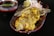 A dish featuring golden-brown grilled pieces of chicken, garnished with thin strands that resemble fried vermicelli, placed in a decorative oval plate. Accompanying the dish is a small bowl of green sauce, a wedge of lemon, and slices of onion placed on the side.