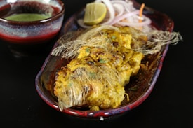 A dish featuring golden-brown grilled pieces of chicken, garnished with thin strands that resemble fried vermicelli, placed in a decorative oval plate. Accompanying the dish is a small bowl of green sauce, a wedge of lemon, and slices of onion placed on the side.