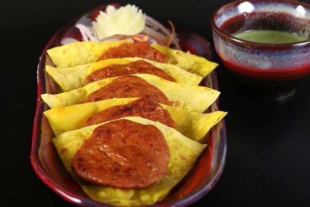 A serving dish contains several yellow chapatis folded over slices of spicy sausage, topped with garnish such as carrots and an onion flower. A small bowl of green chutney is placed beside the dish.