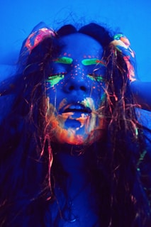 A person with long hair is covered in neon paint that glows under ultraviolet light. The paint is applied artistically across the face, highlighting features like eyebrows and lips with bright colors such as green, orange, and pink. The lighting creates a dramatic and surreal effect.