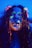 A person with long hair is covered in neon paint that glows under ultraviolet light. The paint is applied artistically across the face, highlighting features like eyebrows and lips with bright colors such as green, orange, and pink. The lighting creates a dramatic and surreal effect.