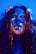 A person with long hair is covered in neon paint that glows under ultraviolet light. The paint is applied artistically across the face, highlighting features like eyebrows and lips with bright colors such as green, orange, and pink. The lighting creates a dramatic and surreal effect.