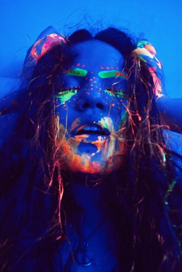 A person with long hair is covered in neon paint that glows under ultraviolet light. The paint is applied artistically across the face, highlighting features like eyebrows and lips with bright colors such as green, orange, and pink. The lighting creates a dramatic and surreal effect.
