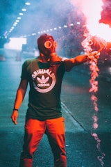 A person wearing a gas mask is holding a lit flare that emits bright red smoke. The background features a dimly lit tunnel with a series of lights creating a dramatic perspective. The individual is dressed in a green T-shirt with a logo and red pants.