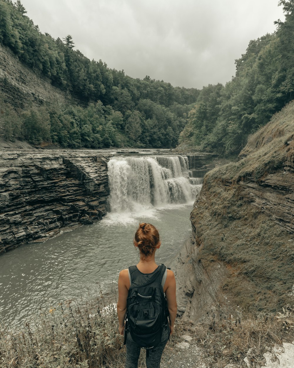 Mujer en camiseta sin mangas negra sentada en la roca cerca de las cascadas durante el día