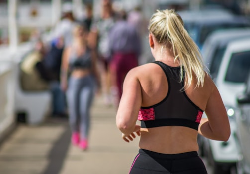 A person with blonde hair is jogging on a pathway in a sporty black and pink outfit. The scene is outdoors, likely in an urban area, with several cars parked to the side. Another person is visible in the background, also dressed in athletic wear.