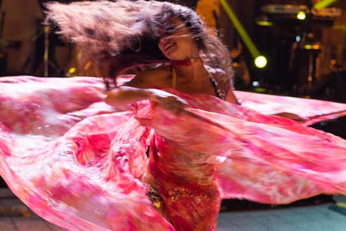 A dancer is captured mid-performance, enveloped in flowing pink and red fabric that appears blurred from motion. Her long hair is flying through the air, creating dynamic movement. The background includes dimly lit stage elements with spotlights.