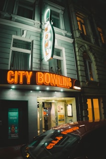A neon-lit bowling alley entrance is prominently featuring a large sign with an illuminated pin design and the words 'City Bowling'. The entrance is surrounded by a classic urban building facade, and the warm glow from the signage creates a cozy, inviting atmosphere.