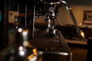 A close-up view of a coffee machine with metallic components, focusing on the spout and the portafilter. The background appears dimly lit with blurred elements, suggesting an indoor cafe setting.