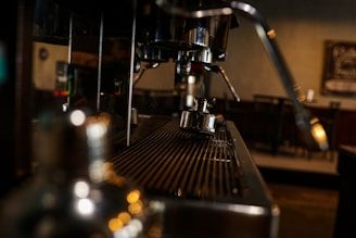 A close-up view of a coffee machine with metallic components, focusing on the spout and the portafilter. The background appears dimly lit with blurred elements, suggesting an indoor cafe setting.