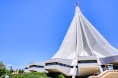 A modern, large architectural structure with a towering, conical design under a bright blue sky. The building has a futuristic appearance with a series of triangular sections converging to a point at the top. Surrounding the base of the structure are several smaller, modular units with large windows.