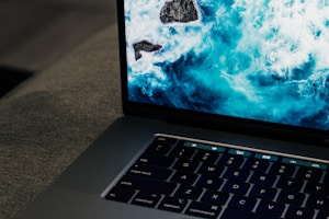 A laptop with a partially visible keyboard featuring illuminated keys. The screen displays a vibrant image of turbulent ocean waves crashing against rocks, creating a dynamic contrast between the serene, dark keyboard and the lively, bright screen.