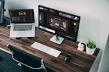 A wooden desk with sleek design features a laptop and a desktop monitor displaying racing game statistics. A wireless keyboard and mouse sit neatly alongside a small plant in a white pot, adding a touch of greenery to the workspace. The chair is positioned under the desk, and the room has a modern, organized aesthetic.