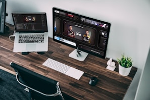 A wooden desk with sleek design features a laptop and a desktop monitor displaying racing game statistics. A wireless keyboard and mouse sit neatly alongside a small plant in a white pot, adding a touch of greenery to the workspace. The chair is positioned under the desk, and the room has a modern, organized aesthetic.