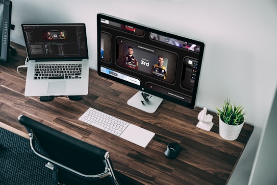 A wooden desk with sleek design features a laptop and a desktop monitor displaying racing game statistics. A wireless keyboard and mouse sit neatly alongside a small plant in a white pot, adding a touch of greenery to the workspace. The chair is positioned under the desk, and the room has a modern, organized aesthetic.