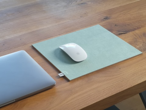A sleek, modern workspace featuring a wooden table with a minimalist design. On the table, there is a minimalist laptop partially visible on the left side. To the right, a white wireless mouse is placed on a grey mouse pad with a simple and clean appearance.