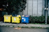 Three large waste bins, two yellow and one blue, are positioned on a sidewalk next to a wall. The bins are overflowing with various types of waste, including plastics and paper. Lush green plants grow alongside the wall behind the bins, adding a natural element to the urban setting.
