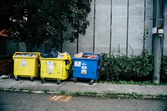 Three large waste bins, two yellow and one blue, are positioned on a sidewalk next to a wall. The bins are overflowing with various types of waste, including plastics and paper. Lush green plants grow alongside the wall behind the bins, adding a natural element to the urban setting.
