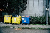 Three large waste bins, two yellow and one blue, are positioned on a sidewalk next to a wall. The bins are overflowing with various types of waste, including plastics and paper. Lush green plants grow alongside the wall behind the bins, adding a natural element to the urban setting.