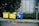 Three large waste bins, two yellow and one blue, are positioned on a sidewalk next to a wall. The bins are overflowing with various types of waste, including plastics and paper. Lush green plants grow alongside the wall behind the bins, adding a natural element to the urban setting.