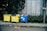 Three large waste bins, two yellow and one blue, are positioned on a sidewalk next to a wall. The bins are overflowing with various types of waste, including plastics and paper. Lush green plants grow alongside the wall behind the bins, adding a natural element to the urban setting.