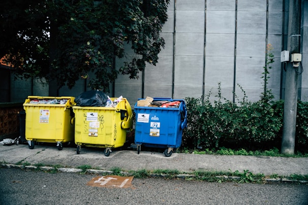 Three large waste bins, two yellow and one blue, are positioned on a sidewalk next to a wall. The bins are overflowing with various types of waste, including plastics and paper. Lush green plants grow alongside the wall behind the bins, adding a natural element to the urban setting.