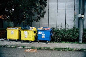Three large waste bins, two yellow and one blue, are positioned on a sidewalk next to a wall. The bins are overflowing with various types of waste, including plastics and paper. Lush green plants grow alongside the wall behind the bins, adding a natural element to the urban setting.