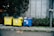 Three large waste bins, two yellow and one blue, are positioned on a sidewalk next to a wall. The bins are overflowing with various types of waste, including plastics and paper. Lush green plants grow alongside the wall behind the bins, adding a natural element to the urban setting.