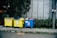Three large waste bins, two yellow and one blue, are positioned on a sidewalk next to a wall. The bins are overflowing with various types of waste, including plastics and paper. Lush green plants grow alongside the wall behind the bins, adding a natural element to the urban setting.