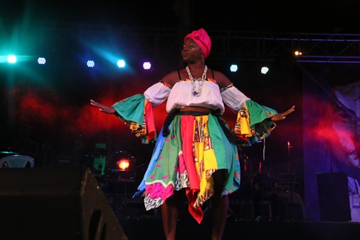 A performer on stage in a colorful costume with bright lights in the background. The attire includes a multicolored skirt, a white top with vibrant sleeves, and a pink head wrap. The scene is dramatic with a backdrop of red and blue lights adding to the atmosphere.