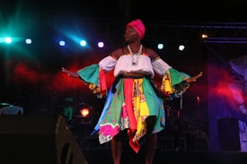 A performer on stage in a colorful costume with bright lights in the background. The attire includes a multicolored skirt, a white top with vibrant sleeves, and a pink head wrap. The scene is dramatic with a backdrop of red and blue lights adding to the atmosphere.