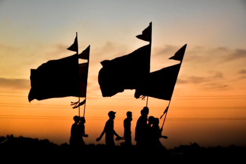 A group of silhouetted people walking, holding large flags against the backdrop of a vivid sunset. The sky is an orange and yellow gradient, with a few scattered clouds. The horizon is lined with dark, indistinguishable shapes, possibly trees or buildings.