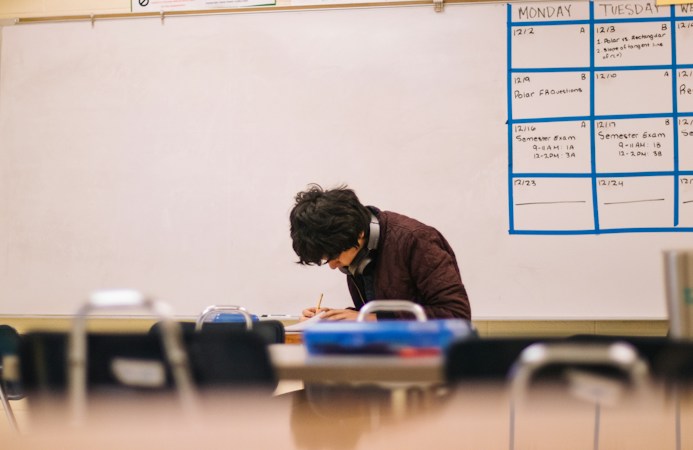 A student writing on paper at a classroom desk during a test session