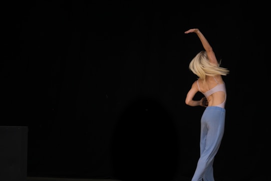 A dancer with long blonde hair gracefully moves against a dark background, wearing a light-colored top and loose blue pants. The pose suggests fluidity and elegance, with one arm raised above the head.