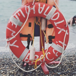 A bright red lifebuoy is positioned on a pebble beach, accompanied by a life jacket. There's a rope attached to both the lifebuoy and jacket. The sea is visible in the background with some waves, and a person is seen partially in the water to the right.