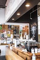 The image depicts the interior of a coffee shop with a wooden counter decorated with various coffee-making equipment. Behind the counter is a modern espresso machine and several coffee cups. The wall in the background is adorned with numerous posters and notices, suggesting a community-oriented atmosphere. Overhead lighting fixtures provide a warm glow.