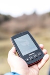 A hand holds a portable GPS device displaying a map. The device shows navigation buttons and the brand name 'TwoNav'. The background is blurred, suggesting outdoor surroundings.