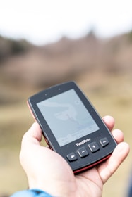 A hand holds a portable GPS device displaying a map. The device shows navigation buttons and the brand name 'TwoNav'. The background is blurred, suggesting outdoor surroundings.