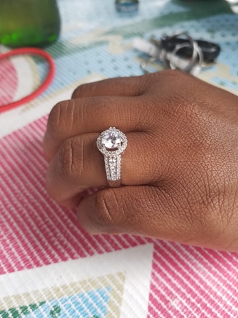A hand adorned with an ornate diamond ring featuring a large central gemstone surrounded by smaller diamonds on a double band. The background includes a patterned tablecloth and blurred objects like keys.