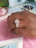 A hand adorned with an ornate diamond ring featuring a large central gemstone surrounded by smaller diamonds on a double band. The background includes a patterned tablecloth and blurred objects like keys.