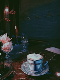 A well-decorated coffee cup and saucer sit on a dark marble table in an ambient, dimly lit setting. The table is adorned with decorative crystal glasses and pink flowers. In the background, a blue-gray brick wall is visible with several empty coat hangers.