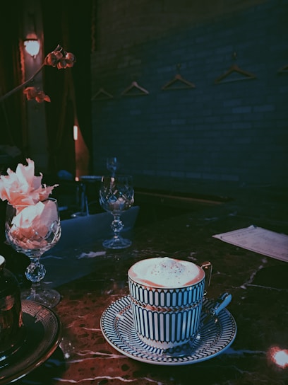 A well-decorated coffee cup and saucer sit on a dark marble table in an ambient, dimly lit setting. The table is adorned with decorative crystal glasses and pink flowers. In the background, a blue-gray brick wall is visible with several empty coat hangers.