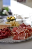 A close-up of a charcuterie board arranged with thin slices of cured meats, cheese cubes, a soft cheese wheel, and some olives in a bowl. The background appears to be a dining setting with bright natural light coming from a window.